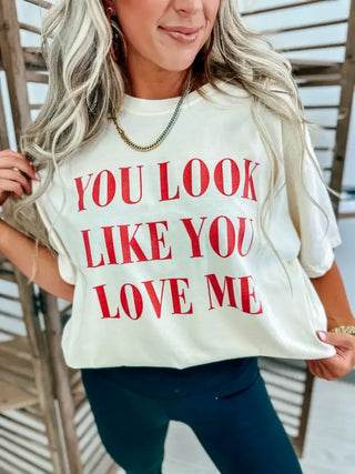 A person with long wavy hair is wearing the "You Look Like You Love Me" Ivory Graphic Tee by Bird & Co Wholesale Tees. They stand before wooden slats, resembling a country love ballad, and are accessorized with a necklace and earrings.