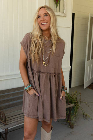 A woman with long blonde hair stands smiling, wearing a Meadow Subtle Escape Textured Dress from Three Bird Nest. The dress is made of ribbed fabric and features a gathered waist with boho western vibes. She accessorizes with multiple layered necklaces and colorful bracelets. One hand rests in a pocket of her dress as she stands in a light-filled, cozy room.