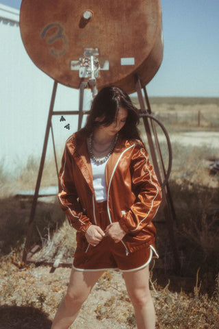 A woman stands outdoors beside an old rusted tank, dressed in a shiny rust brown WYO WEST SET Jacket and Short Satin from 2FLY, complete with white stripes. She pairs it with a white top and layered necklaces. The background showcases dry grass stretching toward a distant horizon under a clear blue sky.