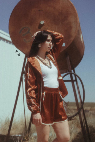 A woman stands outdoors in front of a large rusted metal tank. She is wearing a white top and the shiny red WYO WEST SET Jacket and Short Satin by 2FLY, with one hand resting on her head. She has long dark hair and appears to be looking into the distance. The sky is clear and blue.