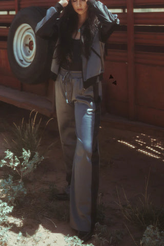 A woman wearing a 2 Fly MARFA MOB Jacket and Pant Set stands in front of a red truck. Her hands are holding her long dark hair back. She is on dirt ground with some green plants around her. The lighting creates a contrast between shadow and sunlight.