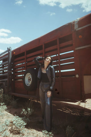 A woman with short hair stands in front of a weathered red wooden truck, striking a pose with one hand on her head and the other on her hip. She confidently sports the MARFA MOB Jacket and Pant Set by 2 Fly under a bright, partly cloudy sky in a dry, desert-like setting.