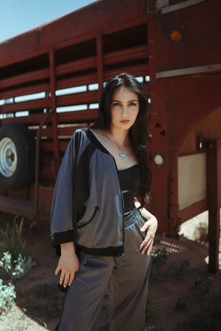 A woman with long dark hair stands confidently in front of a red truck in an outdoor setting. She is wearing a stylish MARFA MOB Jacket and Pant Set by 2 Fly with a black top underneath, complemented by a turquoise necklace. The sun casts soft shadows around her, highlighting her relaxed pose.