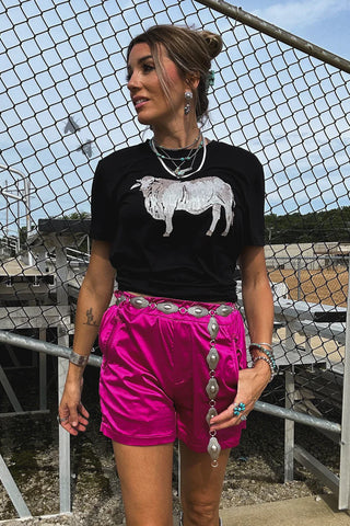 A person with braided hair stands outside in front of a metal fence, wearing a black shirt featuring a sheep graphic and vibrant pink Layken Cowboy Tuxedo Shorts from 2 Fly, secured with a decorative belt. They have various accessories including necklaces, earrings, and bracelets.