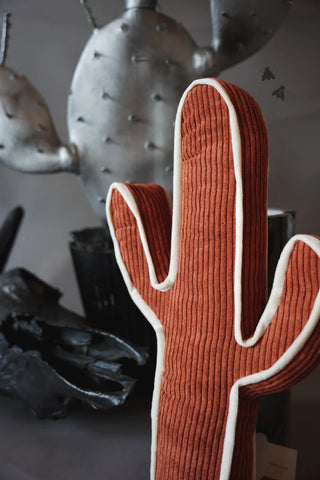 In the foreground, the KICK A CACTUS PILLOW from Western Edge Boutique, a plush rust-colored cushion with white edges, introduces a touch of western style to your home decor. Behind it stands a striking metallic cactus sculpture in silver tones, with several black flies perched on its surface.