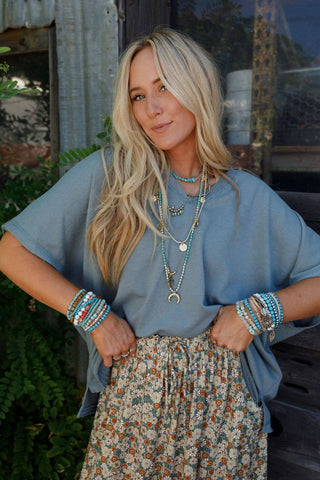 A woman with long blonde hair stands with her hands on her hips, donning the Payton Top by Three Bird Nest, which is a loose blue-green shirt. She pairs it stylishly with a floral skirt and numerous colorful beaded bracelets and necklaces. Her bohemian style outfit pops against the background of greenery and a rustic, corrugated metal wall.