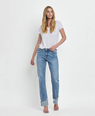 A person with long blonde hair is wearing a white T-shirt, blue Dezie Mid Rise Cuffed Straight Jeans from Lovervet by VERVET, featuring frayed hems and soft stretch denim in sizes 1-24, and silver high-heeled sandals. They are standing against a plain white background.