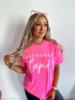 A woman with long blonde hair in a Bird & Co Wholesale Tees "Not Today Cupid" Neon Pink Graphic Tee stands against a white background, holding her hair and slightly smiling. Her sunglasses resting on her head capture the Anti-Valentine vibe perfectly.