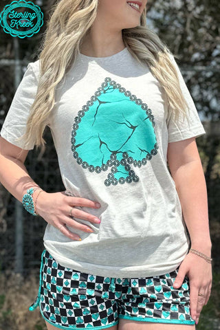 A woman stands outdoors wearing the Sterling Kreek My Lucky Day Tee, which features a large marble turquoise spade symbol. She pairs it with patterned shorts in black, white, and turquoise hues. A turquoise bracelet adorns her wrist while her long blonde hair flows freely against a backdrop of blurred foliage.