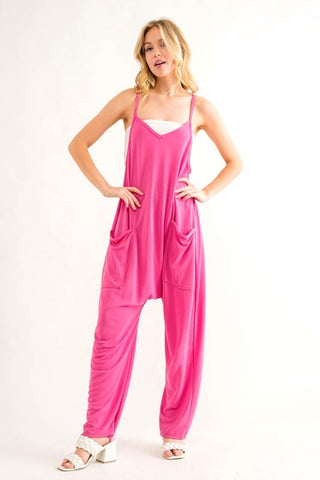 A woman standing against a white background wears the Fashion District Hot Pink Henley Harem Cami Relaxed Jumpsuit featuring spaghetti straps and loose-fitting pants. She pairs it with white sandals and has her hands in her pockets. Her blonde hair is styled in loose waves, complementing her relaxed jumpsuit look.