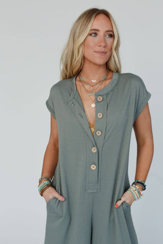 A woman with long blonde hair is seen wearing a Three Bird Nest Logan Wide Leg Jumpsuit in light olive as she stands against a plain white background. Her hands rest in the pockets of the jumpsuit, and she is adorned with multiple layered necklaces and bracelets. She gazes slightly to the side with a subtle smile on her face.