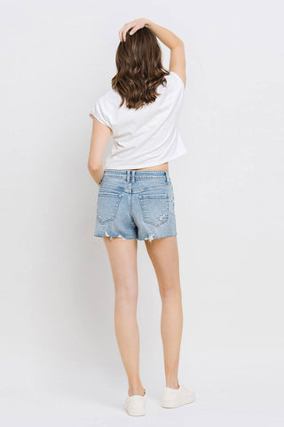 A person with shoulder-length brown hair is facing away from the camera, wearing a white T-shirt and Kylie Mid Rise Raw Distressed Hem shorts in light blue comfortable stretch denim by VERVET by FLYING MONKEY. The outfit, perfect for all sizes, is completed with white sneakers. One arm is raised, touching the back of their head against the plain white background.