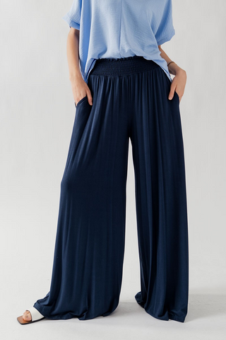 A person wearing chic and sophisticated Trend Notes Keren Bamboo Rayon Wide Leg Palazzo Pants in navy blue with an elastic waistband and a light blue top. The person has their hands in the pockets of the flowy pants and is standing against a plain white background. The person's head and feet are not visible in the image.