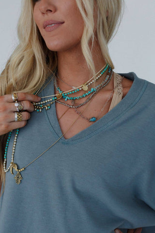 A person with long blonde hair wearing the Payton Top by Three Bird Nest, a blue v-neck made of stretchy knit fabric, showcases multiple layered necklaces adorned with turquoise beads and a hamsa hand pendant, encapsulating the bohemian style. They also feature several rings on their fingers against a plain gray background.