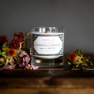 A lit Coastal Cowgirl Candle 8 oz. from R. Rebellion, encased in a clear glass holder with a label that reads "R. Rebellion Coastal Cowgirl, made in the West." The candle emits musk notes and oceanic salt, placed on a wooden surface with dried flowers scattered around it against a dark gray background.