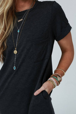 Close-up of a person wearing the On The Daily Pocket Tee Dress in charcoal from Three Bird Nest and several layered necklaces with pendants. The person has long, wavy blonde hair and is sporting multiple colorful beaded bracelets on one wrist, with the hand tucked into their pocket. The background is plain white.