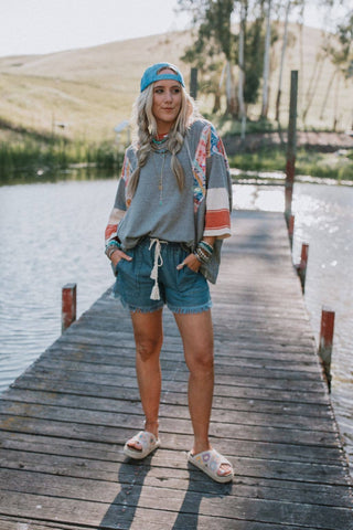 A woman stands on a wooden dock over a lake, wearing a colorful cap, grey oversized shirt with patterned sleeves, Three Bird Nest's "Meet The Moment Drawstring Shorts" in Dark Wash, and sandals. The background includes calm water and serene, grassy hills. The sunlight casts a soft glow over the scene.