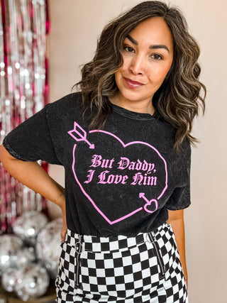 Wearing Mugsby's But Daddy I Love Him Valentine's Shirt, a person with wavy hair stands against shiny streamers. The vintage-inspired black graphic tee has a pink heart and pairs perfectly with checkered pants, evoking the charm of an old country song.