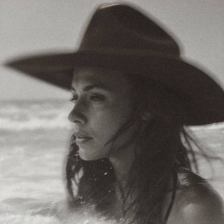 A monochromatic image depicts a person with wet hair, donning a large-brimmed hat, and gazing to the side. The background suggests an ocean or sea with gentle waves, evoking a sense of calm and introspection akin to the unique and playful scent of R. Rebellion's Coastal Cowgirl Perfume 50 ml.