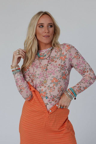 A blonde woman wearing the "Made You Blush Floral Top - Pink Floral" by Three Bird Nest and an orange overall skirt looks to the side. She accessorizes with multiple bracelets, rings, and necklaces. The background is plain white.