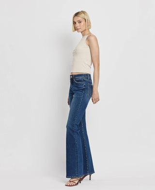 A woman with a blonde bob haircut stands sideways wearing a sleeveless white top, Kip Mid Rise Relaxed Bootcut Jeans by Lovervet by VERVET made from comfort stretch denim, and black high-heeled sandals against a plain white background.