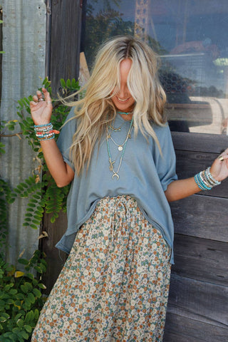 A person with long blonde hair wearing the Payton Top by Three Bird Nest, paired with a floral maxi skirt and adorned with various colorful bracelets and necklaces that epitomize bohemian style, stands in front of a rustic wooden door surrounded by greenery. They are smiling with their head slightly bowed, holding their hair.