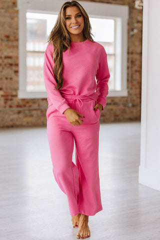 A woman stands smiling in a bright room with brick walls and large windows, wearing the Stockton Textured Set by Liam & Company. This pink ensemble features a matching sweater and wide-leg pants, with exquisite textures complementing her barefoot look on the light-colored floor.