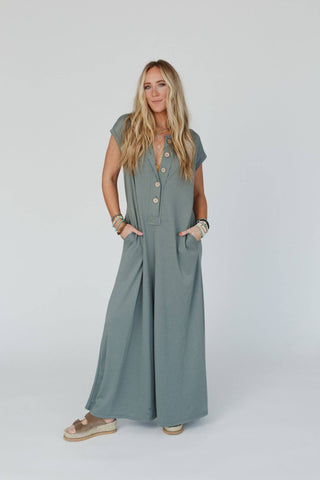 A woman stands against a plain white background wearing a loose, floor-length Logan Wide Leg Jumpsuit - Light Olive by Three Bird Nest, featuring short sleeves and buttons down the front. She has long, wavy blonde hair and is accessorized with various bracelets and necklaces. She completes her look with platform sandals.