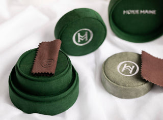 A close-up of two green velvet jewelry boxes, one open and one closed. Both feature a monogram with the letters "KM." Inside the open box, a brown polishing cloth showcases the Haisley French Twist Chain Necklace 925: 2mm from Moxie Maine. The background is a soft white fabric.