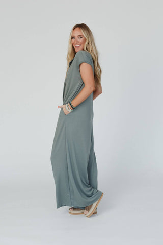 A woman with long blonde hair smiles while posing. She is wearing the Logan Wide Leg Jumpsuit in light olive by Three Bird Nest, several bracelets on her wrist, and beige wedge sandals. She stands against a plain white background with her left hand in her jumpsuit pocket.