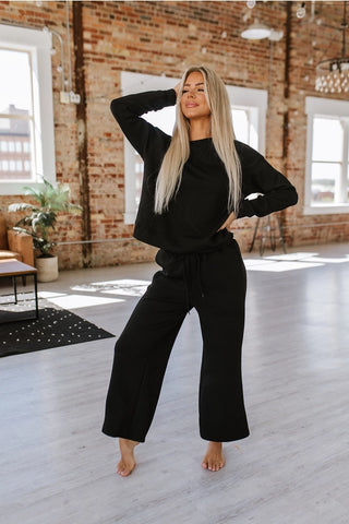 In a sunlit room with exposed brick walls and large windows, a woman with long blonde hair poses gracefully. She is dressed in the Alex Black Lounge Set from LIAM AND CO, a perfect blend of comfort and style that features a sleek black top paired with matching pants. One hand rests on her head while the other is placed on her hip, complementing the minimalistic decor surrounding her.