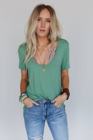 A woman with long, blonde hair is standing against a neutral background. She is wearing the green Moxen Scoop Neck Bralette Tee by Three Bird Nest, featuring a curved hemline. She pairs it with layered necklaces and blue jeans, her hands tucked into her pockets. Her wrist is adorned with several bracelets as she gazes directly at the camera.