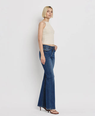 A person with short blond hair is standing against a white background, wearing a sleeveless beige top and Kip Mid Rise Relaxed Bootcut Jeans by Lovervet by VERVET. They are also wearing brown strappy sandals. The person is facing slightly to the left with their hands by their sides.