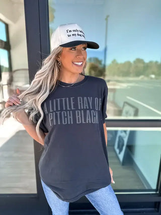 A woman with long hair, wearing a cap and a Bird & Co Wholesale Little Ray Of Black Black Graphic Tee (Large), stands outside near a glass door. She's holding her hair and smiling, set against a blurred backdrop of trees and parking.