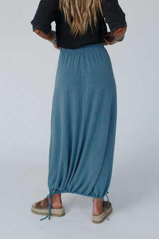 A person with long hair is standing with their back to the camera, wearing a black top and the Rylie Drawstring Maxi Skirt in teal by Three Bird Nest that reaches their ankles. This boho western wardrobe staple features an elastic drawstring waistline and hem, creating a gathered effect. They are also wearing brown sandals.