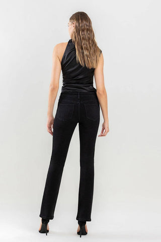 A person is standing with their back to the camera, wearing a sleeveless black top and Lovervet by VERVET's Halston Mid Rise Bootcut Jeans—a true wardrobe must-have. Their long, wavy hair cascades down their back. The individual also sports black heels against the plain light grey background.