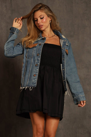 In front of a textured gray backdrop, a woman poses wearing Blue Buttercup's Ryder Aztec Print with Faux Suede Denim Jacket over a strapless black dress. Her long, wavy hair cascades as she looks down, adjusting her jacket with one hand and gently touching her hair with the other.