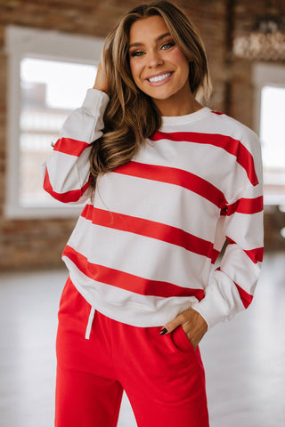 A woman smiles indoors, wearing the Callahan Striped Pullover and Joggers Set by Liam & Company. Her long hair frames her face as she touches her head, while large windows and soft lighting enhance the cozy vibe of her outfit.