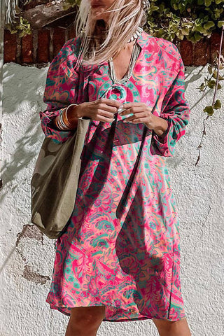 A person with long hair stands against a white wall, wearing a vibrant Retro Paisley Tab Sleeve Dress from Pretty Bash featuring pink, green, and blue paisley patterns. They are accessorized with necklaces, bangles, and rings, and carry a large olive green bag over one shoulder.