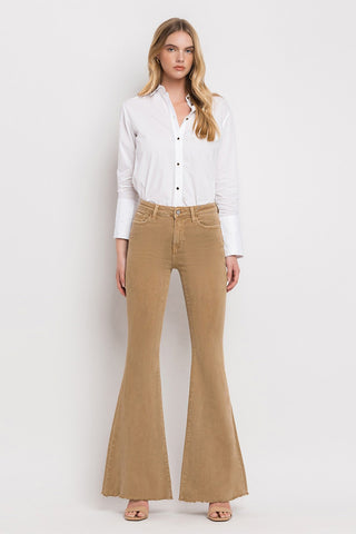 A woman stands against a plain white background wearing a white button-up shirt with the sleeves rolled up and high-waisted, olive green Ellis High-Rise Super Flare Jeans from VERVET by Flying Monkey. She has long, wavy hair and is wearing open-toe shoes. She faces forward with a neutral expression.