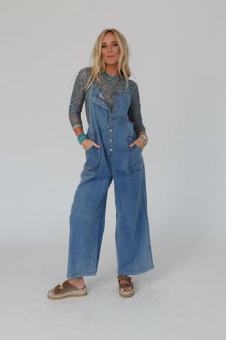 A person with long blonde hair stands confidently against a plain background, wearing the Dock Town Button Up Jumpsuit in denim from Three Bird Nest over a patterned long-sleeve shirt. They have their hands casually in their pockets and wear brown sandals, several bracelets, and rings, embodying the carefree essence of boho style clothing.