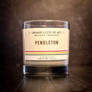 Against a dark background, a Pendleton Candle 8 oz by R. Rebellion in a clear glass jar showcases its label, which reads "Pendleton" and features the text "Somewhere a little out west. Made in the West - Pendleton, Oregon" along with "R. Rebellion, Coconut Soy Candle 8 oz. / 227 g." The candle exudes nostalgic notes reminiscent of 1910 Pendleton Whiskey and spiced pear.