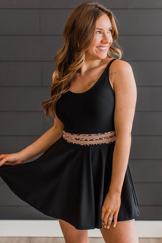 A person with long hair smiles while wearing the "Living On Sunshine Swim Dress- Black & Leopard" from Western Edge Boutique. They hold the hem of the dress, standing in front of a dark paneled wall.