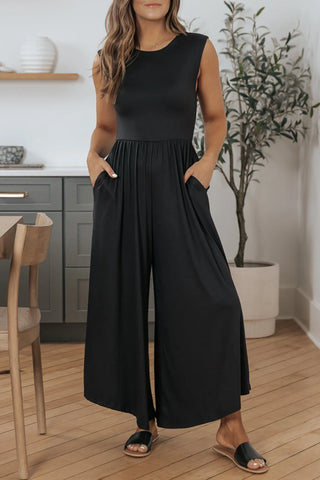 A woman stands in a kitchen wearing the Quincey Black Open Back Wide Leg Jumpsuit from Western Edge Boutique. The sleeveless jumpsuit features a cinched waist and a breathable fabric with side pockets where she rests her hands. She completes her look with black slide sandals. Behind her are gray cabinets, wooden floors, and a potted plant.