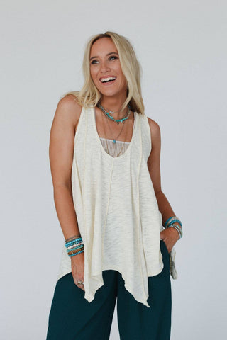 A person with long blonde hair and a cheerful expression is wearing the Elara Top - Neutral, a sleeveless, loose-fitting light beige comfy top by Three Bird Nest. They also have on a blue beaded necklace, several bracelets, and dark green pants. Their hands are in their pockets as they look slightly to the side.