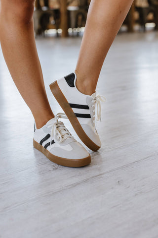 A person stands on a light-colored wooden floor, showcasing the Ness Striped Lace Up Flat Sneakers: White Black from Liam & Company. The fashion sneakers feature black stripes, white laces, and a gum sole. The sleek, casual design of these shoes is highlighted by the visibility of only the person's lower legs and feet.