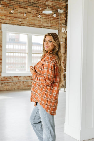 A person with long hair is smiling and looking at the camera while wearing the Palmer Oversized Flannel from Western Edge Boutique in orange plaid, paired with light blue jeans. They are standing in a bright room with brick walls and large windows, reminiscent of a fashionista's closet.