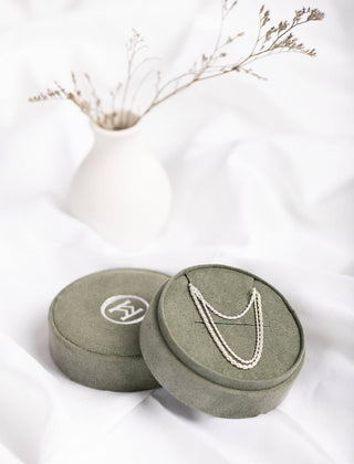 The Haisley French Twist Chain Necklace 925: 2mm by Moxie Maine is delicately displayed on the lid of a closed green velvet jewelry box adorned with a white logo. Both items are elegantly arranged on a soft, white fabric surface. In the background, a simple white vase holds dried flowers.