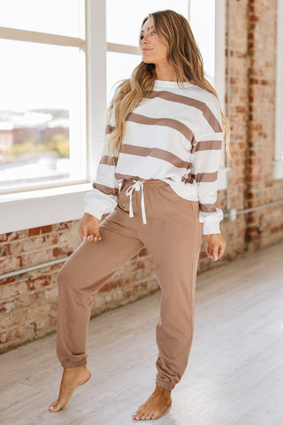 A woman poses by a large window, highlighting the comfortable fit of her Liam & Company Callahan Striped Pullover and Joggers Set in beige and white stripes with brown joggers. She stands barefoot on a light wooden floor against a brick wall with soft natural light streaming in.