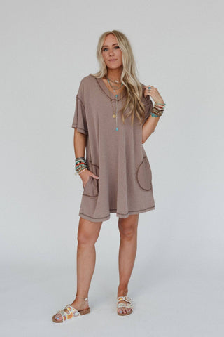 A woman with long blonde hair is wearing a loose, brown Brooklyn Soft Touch Tunic Dress by Three Bird Nest, complemented by multi-colored bracelets. She is also adorned with sandals and various necklaces. The boho-chic dress flows effortlessly as she poses with one hand on her hip and the other resting by her side.